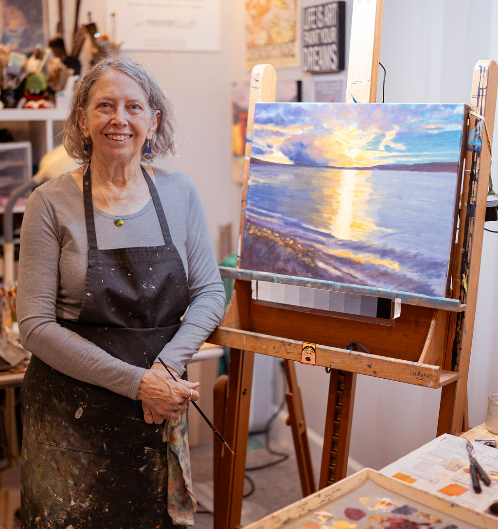 Artist in a studio with a painting of a sunset over water on an easel.